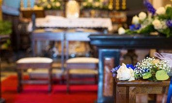 Two flower-decorated chairs face a flower-adorned altar in a chapel, which is surrounded by blurred pews and a red carpet, emphasizing the wedding ceremony setting.